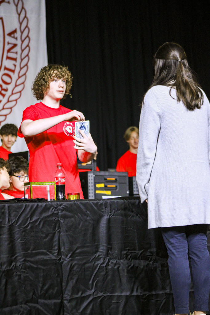 A person in a red shirt holds a card and card deck at a table. A woman watches.