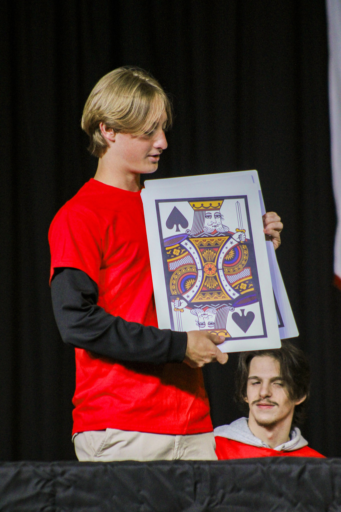 A person holding a card with a face of a king of spades while another person sits below.