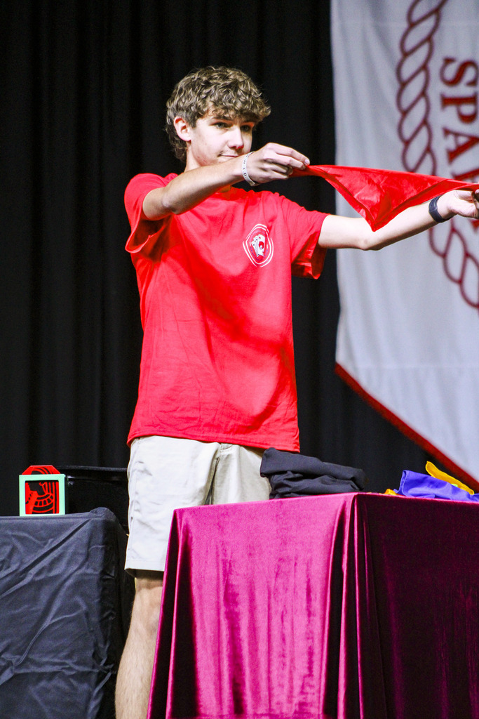A person in a red t-shirt and white shorts performs on stage, holding a red cloth and standing in front of a purple table.