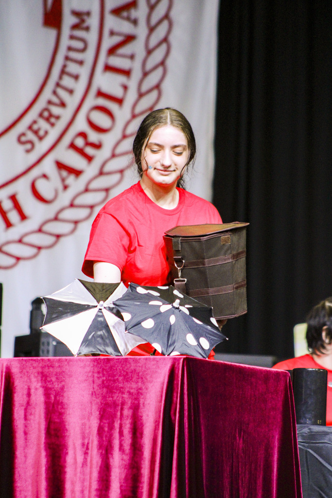 A person in a red shirt stands on a stage with a backdrop of a university logo, holding black objects.