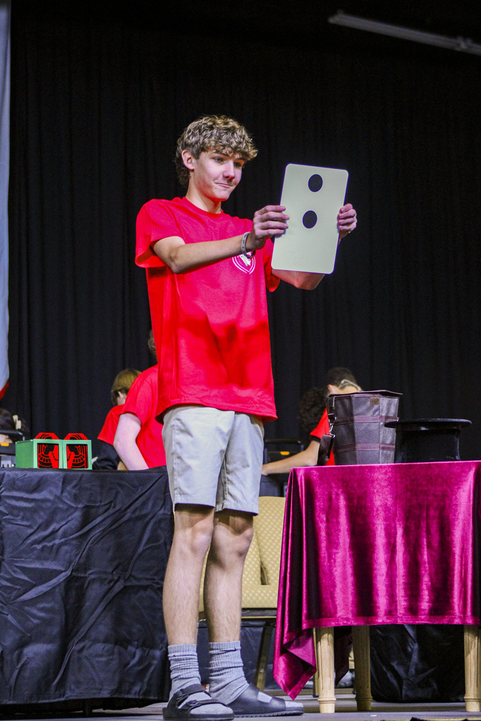 A person in red shirt and khaki shorts stands on stage, holding a large white card with two black circles.