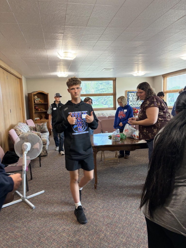A boy stands and walks in a room with people. A fan and furniture are in the background.