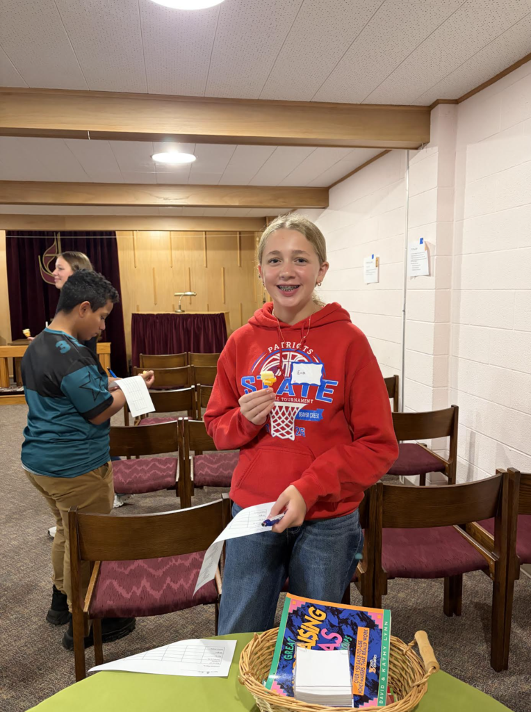 A young person in a red hoodie stands holding a paper and pen. Behind them, a person in a blue shirt reads a paper. Chairs and a table with a basket are in the room.