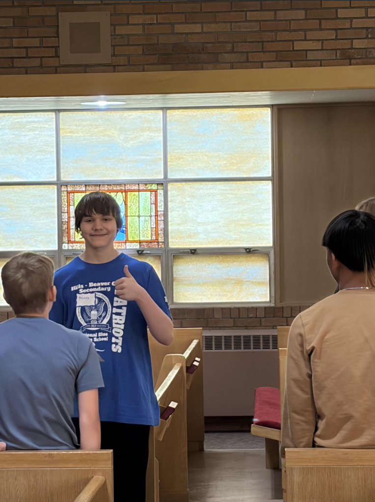 A young boy gives a thumbs up in a church. Behind him, two boys face forward. A stained glass window reflects light.