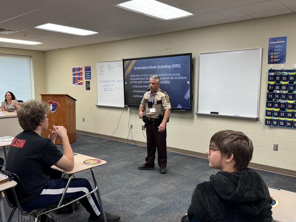 A uniformed officer stands in front of a classroom screen while two students listen attentively.