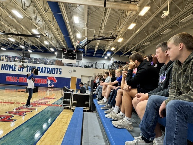A person stands at a basketball court's center, while others sit on bleachers, facing the speaker.