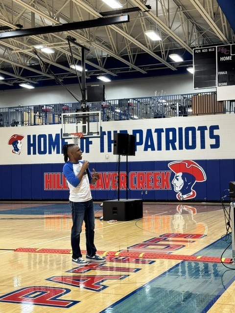 Person stands on basketball court, holding a microphone, in front of a wall that says "Home of the Patriots."
