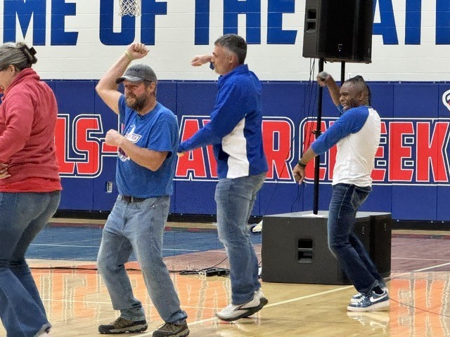 Four adults, in blue and white shirts, dance on a basketball court in front of a wall.