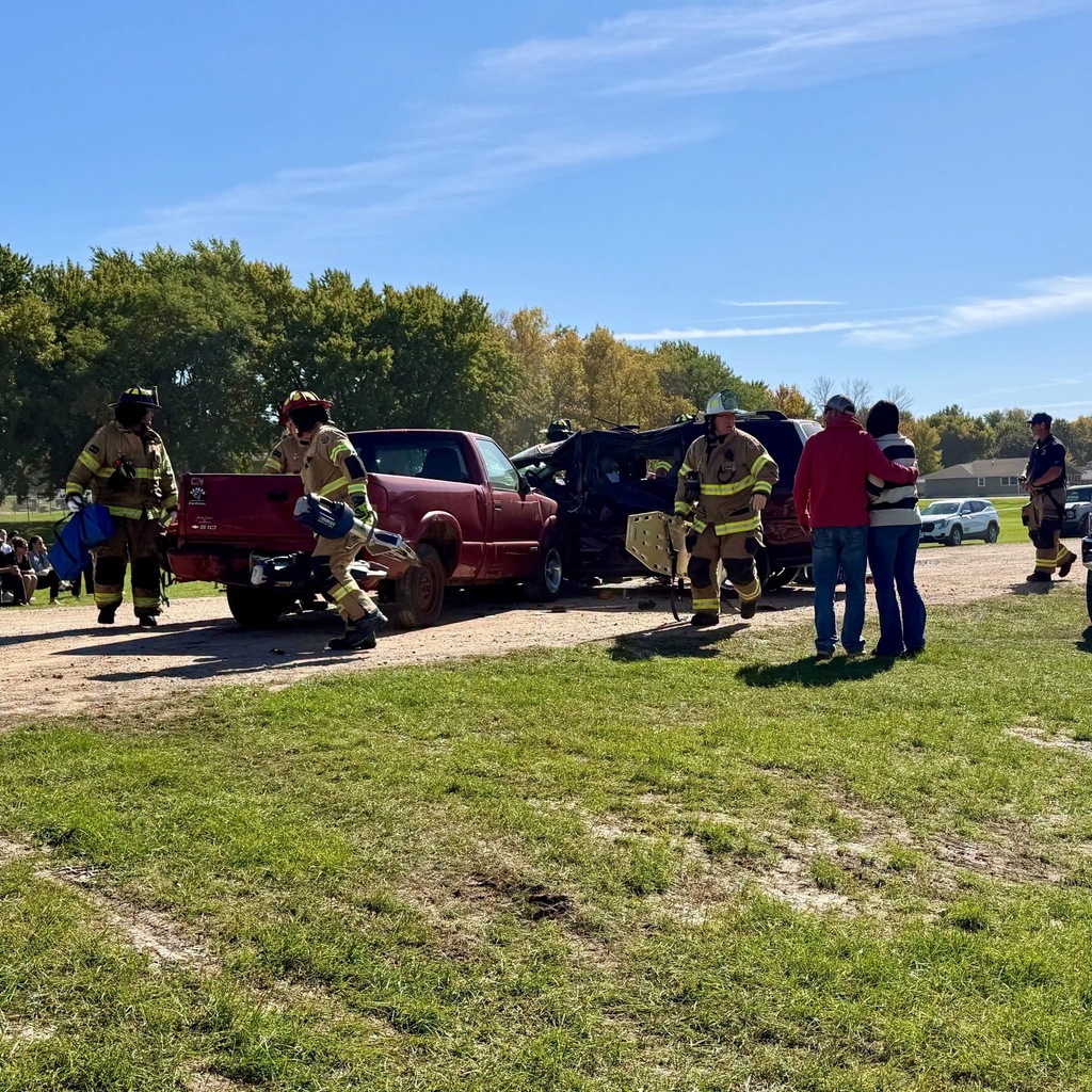 Emergency personnel in gear surround a red pickup truck and black SUV on a gravel road. Trees in the background.
