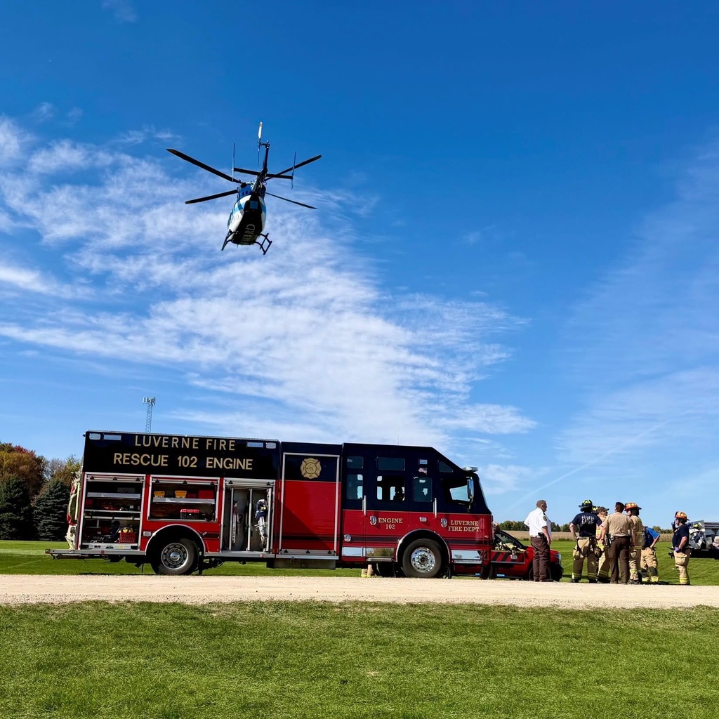 Red fire truck with Rescue 102 on side. Black helicopter above. People around truck. Green grass and clear sky.
