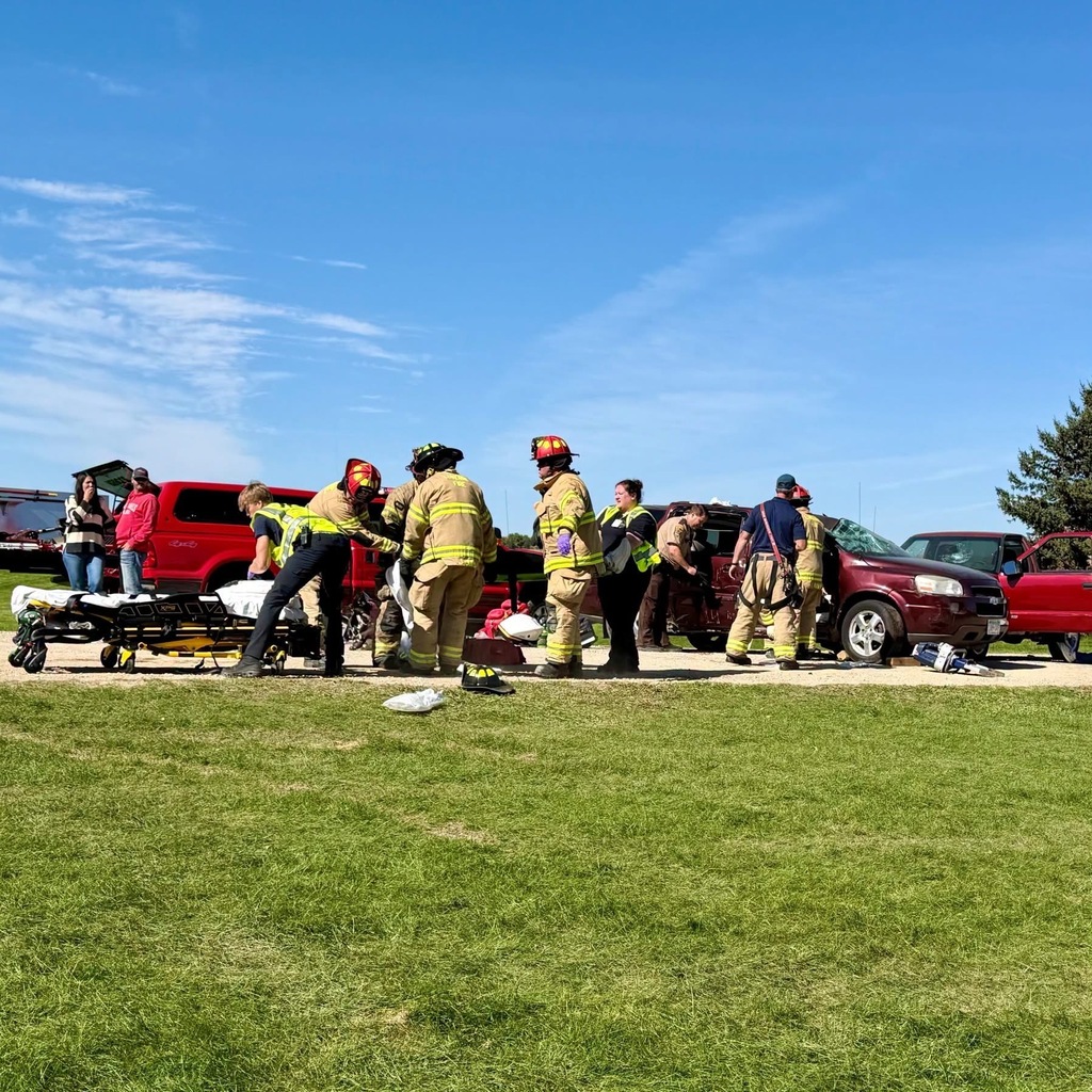 Emergency personnel assist injured people on a grassy field. Cars are parked nearby, and the sky is clear.