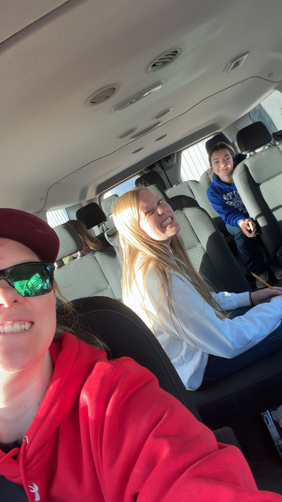 Three people inside a vehicle. A woman in red smiles and wears sunglasses. A girl in white smiles, and another person sits behind her.