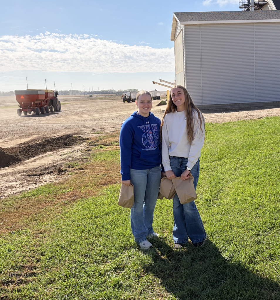 Two people in a field. One wears a blue hoodie, the other a white long-sleeve shirt. Behind them is a structure.