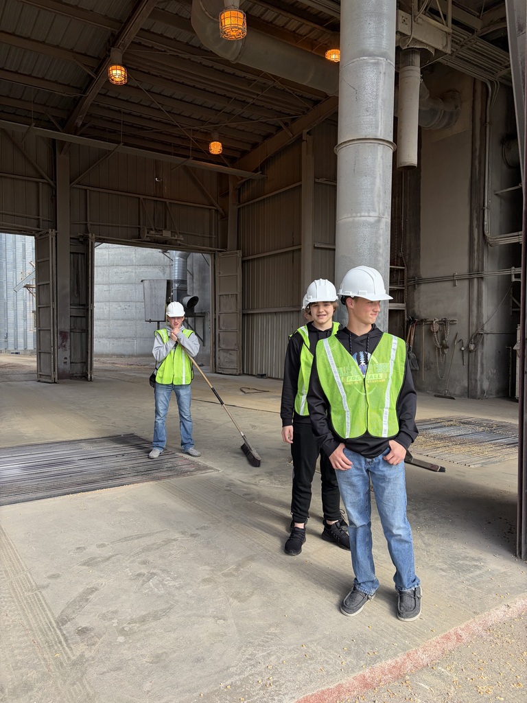Three people in yellow vests and hard hats stand inside an industrial building. One holds a broom.