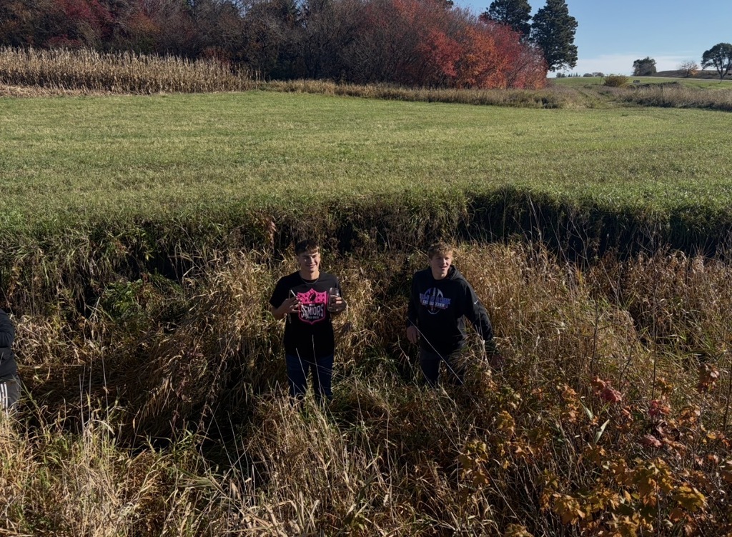 Two students in black shirts stand in a tall grass field with trees and a red-leafed tree.