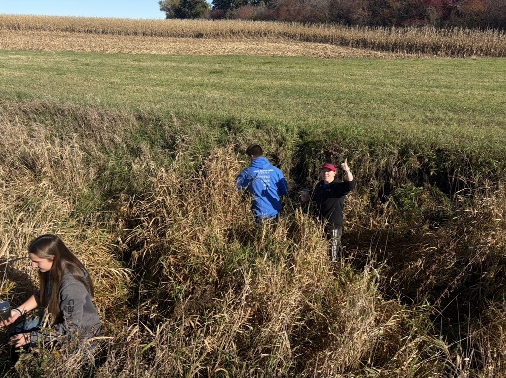 Four individuals in a tall grass field, two with jackets, one with a red hat, one holding a camera.