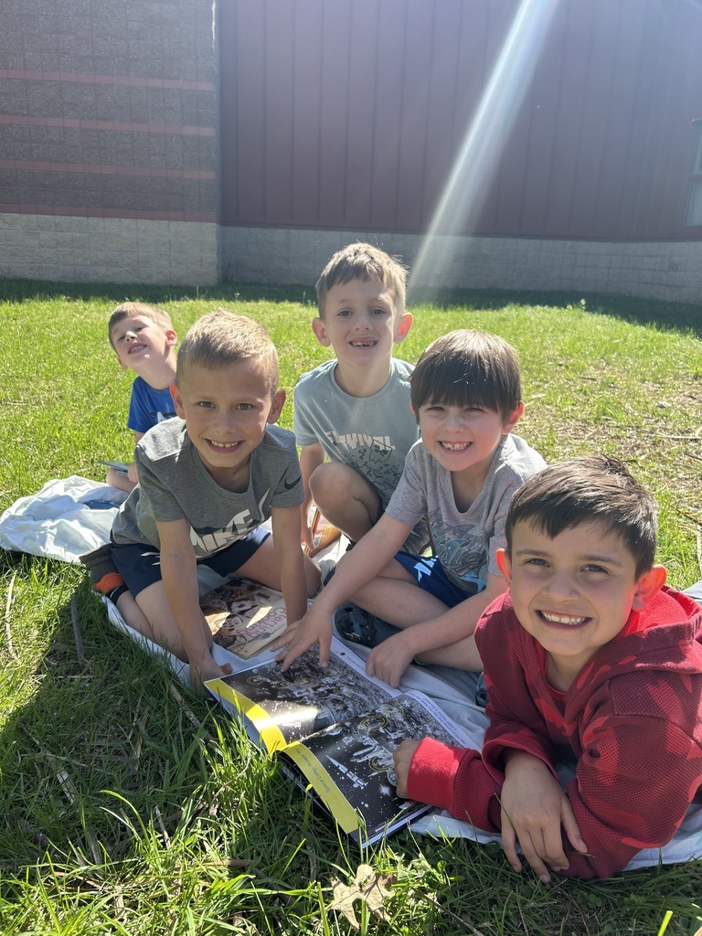 image shows children smiling for a picture as they read books outside on a beautiful day