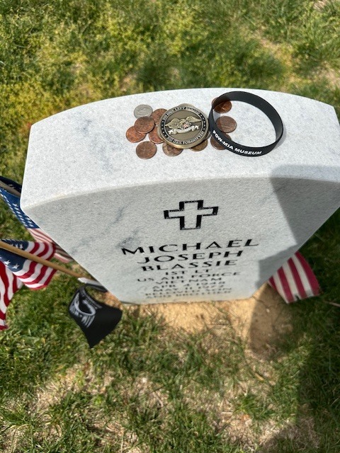 image of a gravesite for 1st Lt. Michael Blassie of the US Air Force. Coins, A Challenge Coin and a bracelet are left at his gravesite. An array of American  flags and a POW/MIA flag are left at the gravesite.
