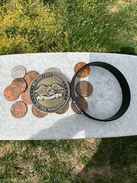 image of a gravesite for 1st Lt. Michael Blassie of the US Air Force. Coins, A Challenge Coin and a bracelet are left at his gravesite. An array of American  flags and a POW/MIA flag are left at the gravesite.