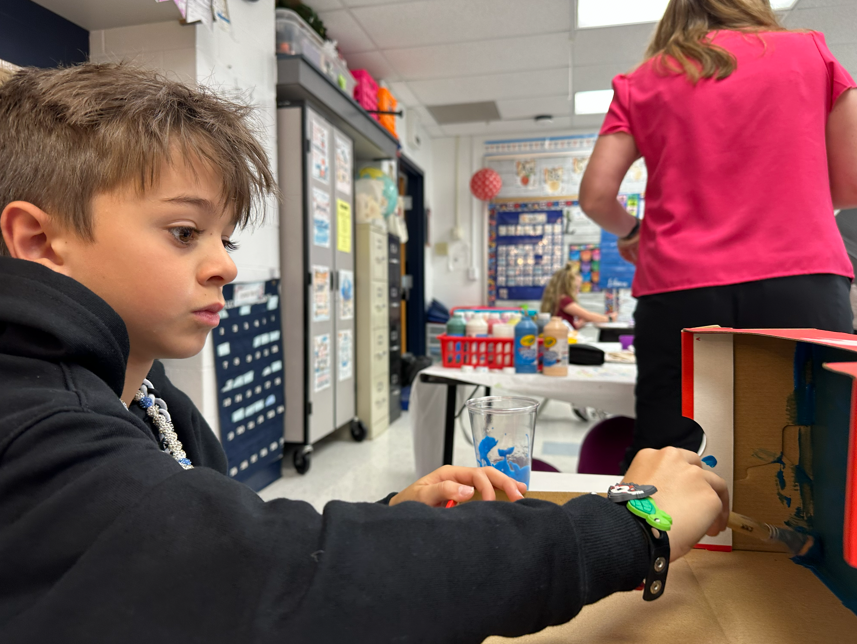 a student paints his shoebox blue as he works on an animal habitat during a class project.