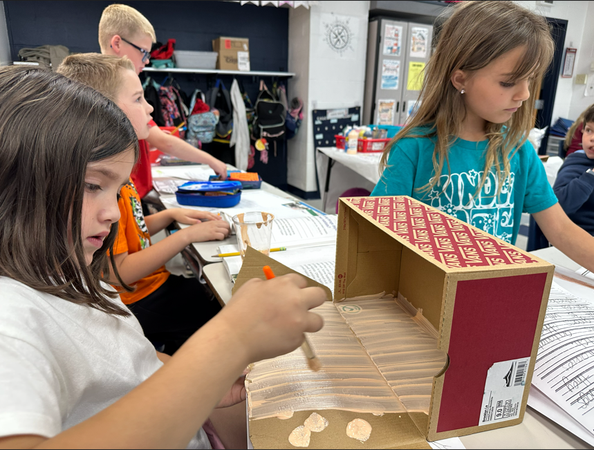 one student in the foreground paints her shoebox animal habitat while a student in the background starts to draw an animal habitat.