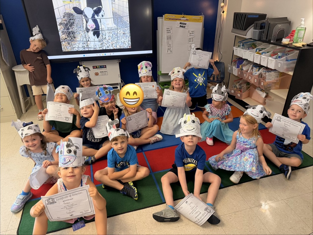 image shows children wearing "cow hats" and smiling at the camera. Mrs. Kansas' class adopted a cow and they receive weekly updates. Part of the image also shows a cow on a TV screen.