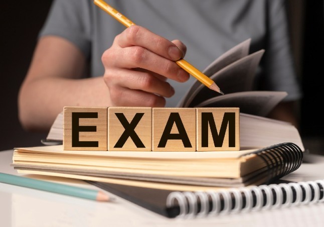 The word Exam in blocks on a notepad with a student holding a pencil behind the word Exam