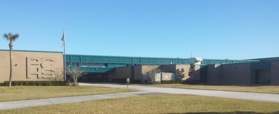 The image contains an exterior view of Plant City High School building on a clear day, with the letters 'PCHS' displayed on the side wall, a flagpole with an American flag, a wide grassy area in the foreground, and a sidewalk leading to the entrance.
