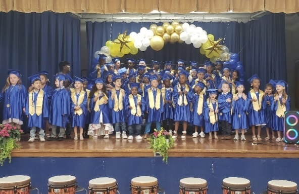 Kindergarten students are standing and smiling on stage at school  in their graduation caps and gowns.