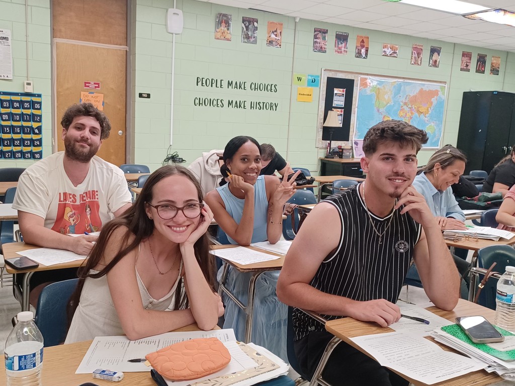 4 students at desks smiling at the camera, while others continue writing