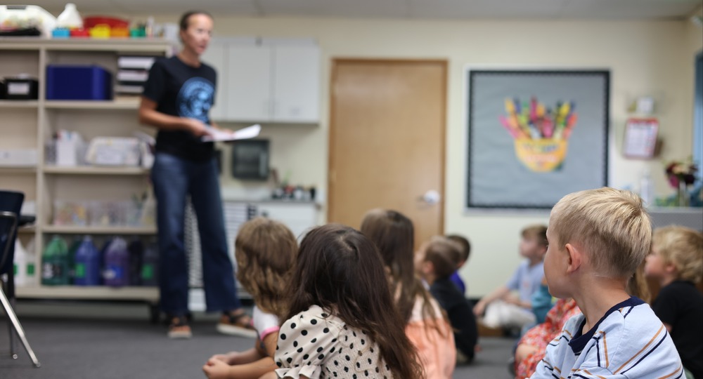 Elementary students sit on the floor and listen to the teacher.