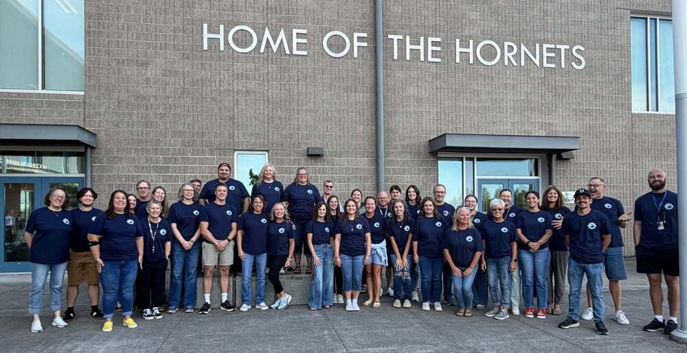 Large group of staff wearing dark blue school spirit shirts in front of a building with lettering that reads Home of the Hornets.