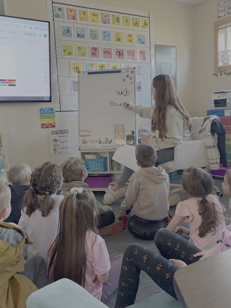 Teacher leads a literacy lesson. Teacher is writing such and thud on a white board while students sit on carpet.