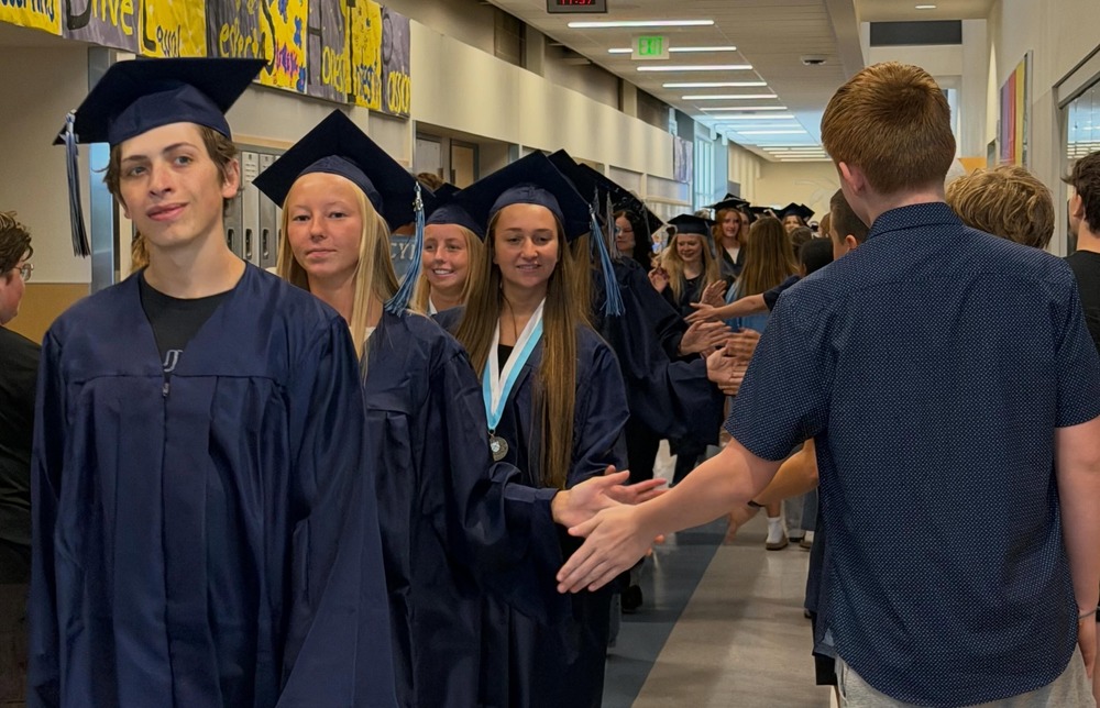 Graduating seniors in caps and gowns high five middle school students in hallway.
