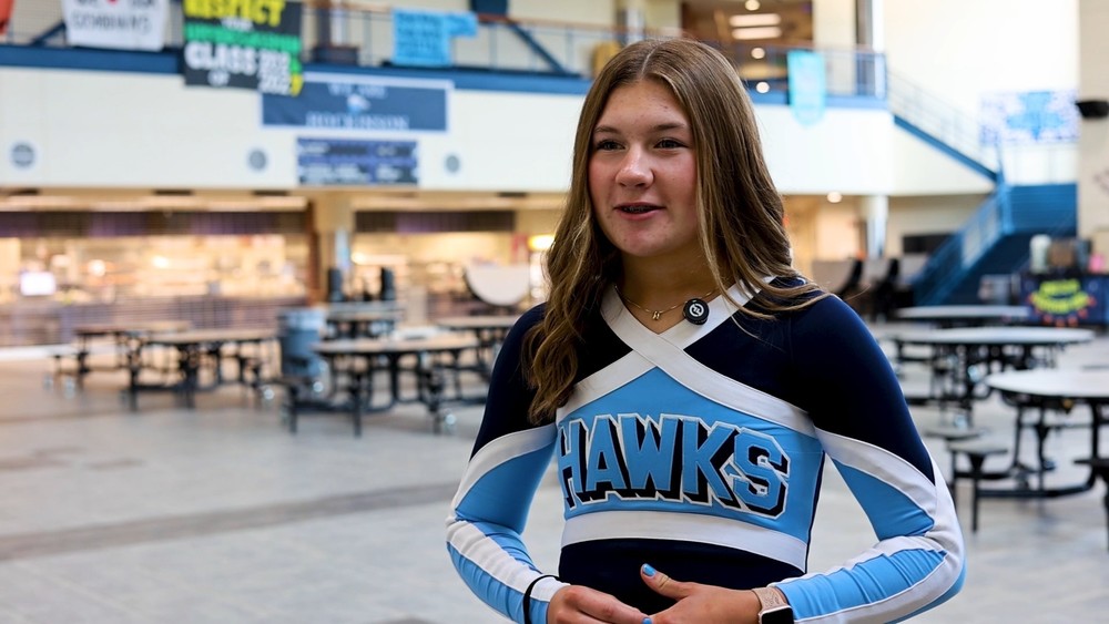 Student in cheer uniform stands in indoor commons area at high school.
