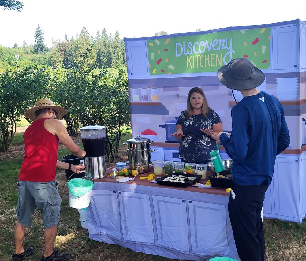 Three people outdoors at a booth titled Discovery Kitchen with fresh food and drink samples on the table.
