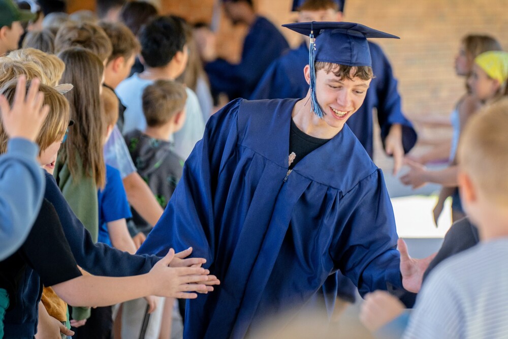 Graduating seniors in caps and gowns high five middle school students in hallway.
