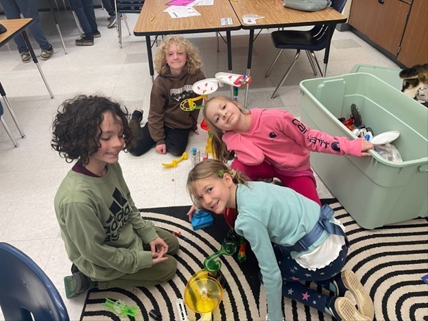 Elementary students sit on ground with marble funnel toys.