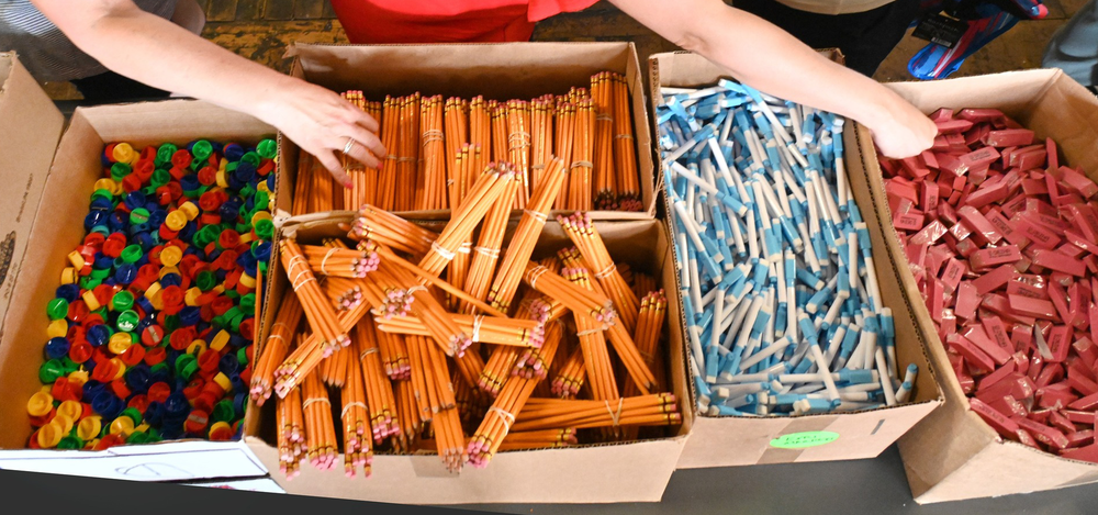 The hands of volunteers above boxes of school supplies, which form a bag packing "assembly line"