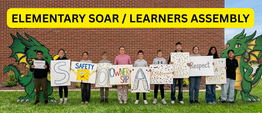 A group of elementary-aged students stands on a grassy lawn in front of a brick school building, holding decorated signs. Large illustrated green dragons flank both sides of the group. A bright yellow banner across the top reads “ELEMENTARY SOAR / LEARNERS ASSEMBLY.” The students’ signs feature the letters and words “S,” “SAFETY,” “O,” “OWNERSHIP,” “A,” “Achievement,” “R,” “Respect,” and “A,” with the final sign also displaying “GOAT.” One student holds a certificate labeled “Congratulations.” The signs are decorated with colorful stars, dots, drawings, and cartoon imagery.