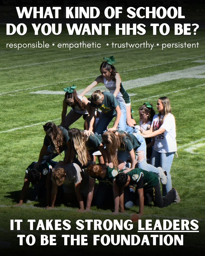 The image shows a group of students engaging in a team-building exercise on a grassy field. The participants are forming a human pyramid, with several students kneeling on the ground to provide support, while others climb on top to form higher layers. The students are dressed in casual attire, some wearing school spirit clothing. Two students on top of the pyramid wear green bows in their hair. A person stands to the side, overseeing the activity, wearing a white shirt with printed text. The field displays white painted lines commonly found on sports fields.