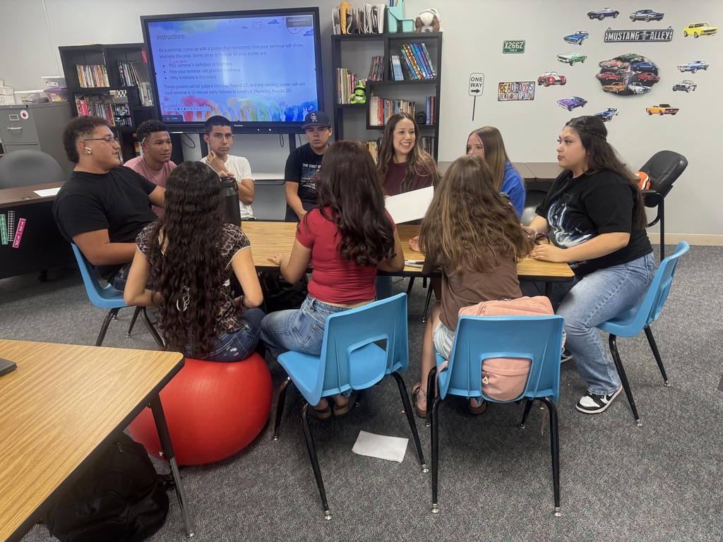 A diverse group of ten students engages in discussion around a circular table in a classroom, with a projection screen displaying project instructions in the background.