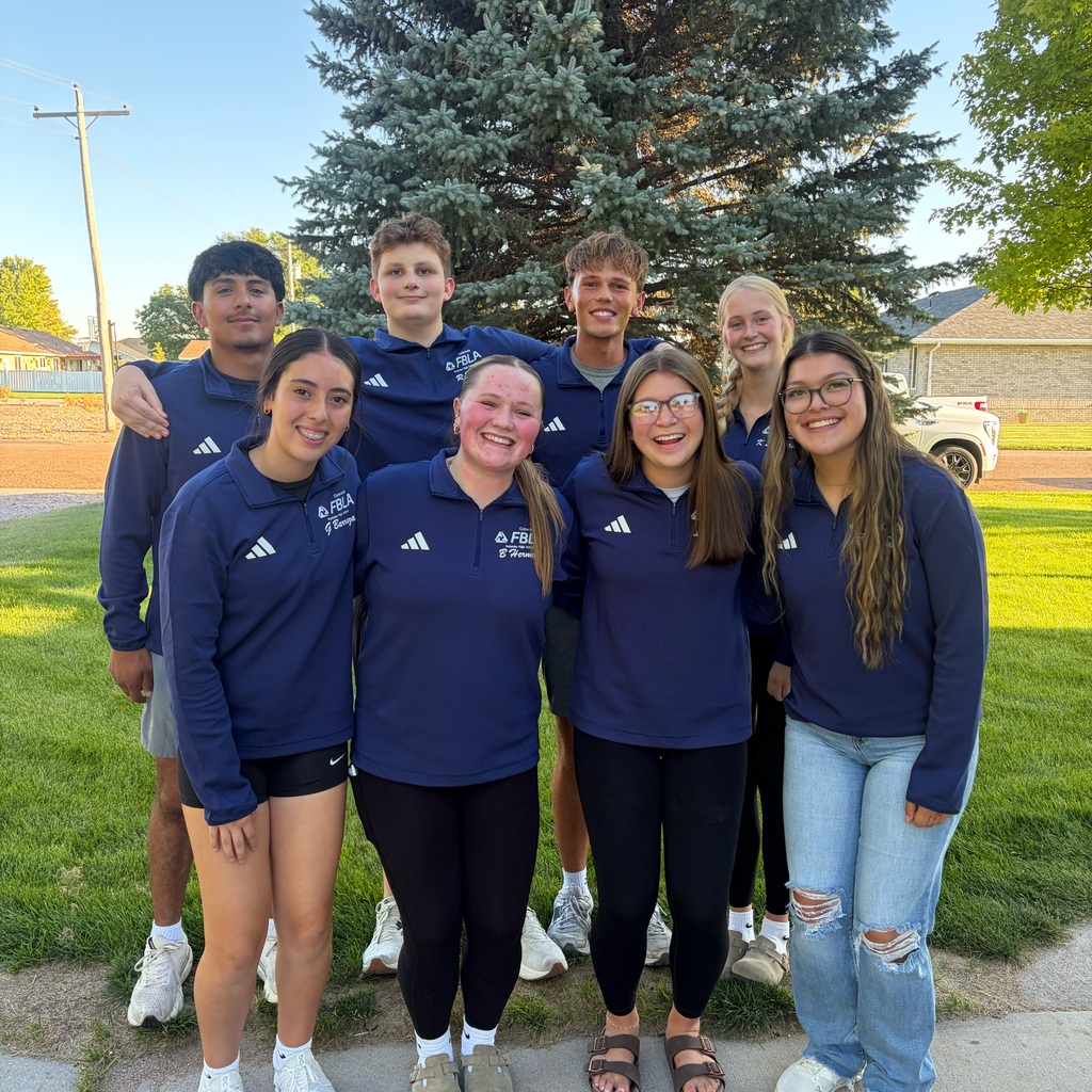 A group of eight young people in matching navy blue shirts smiles together outdoors in front of a green tree and residential area.