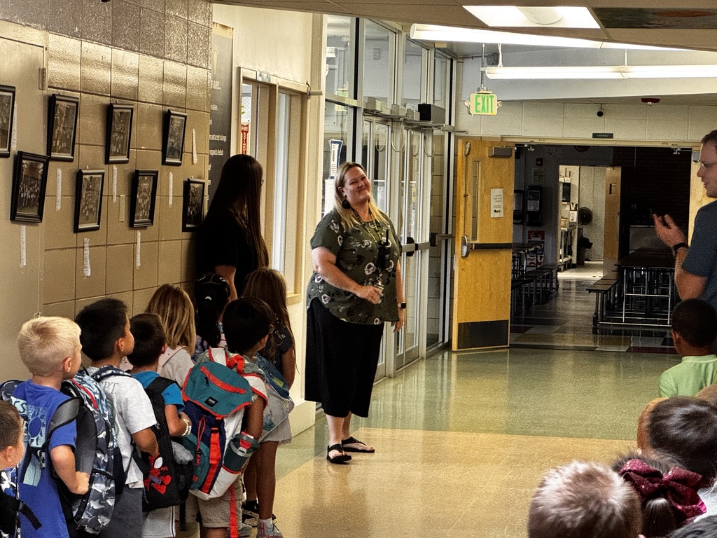Students lined up in the hallway. There is an adult at the front of the line who appears to be very happy.