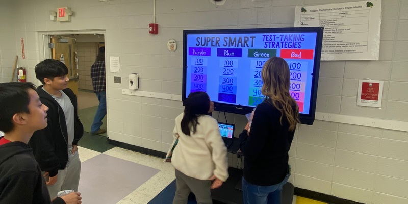 students play a jepordy! style game on a interactive touch screen with a teacher watching.