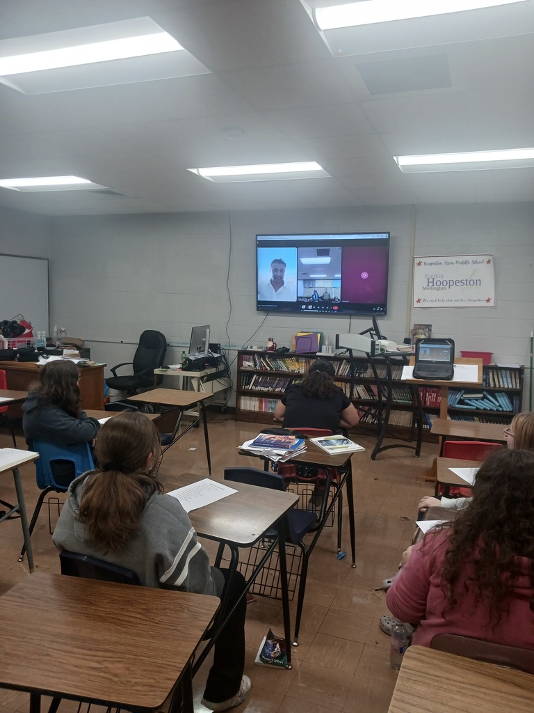 Students sit in desk and look at the screen on the wall as Tyler Jacob Moore joins them for a remote interview.