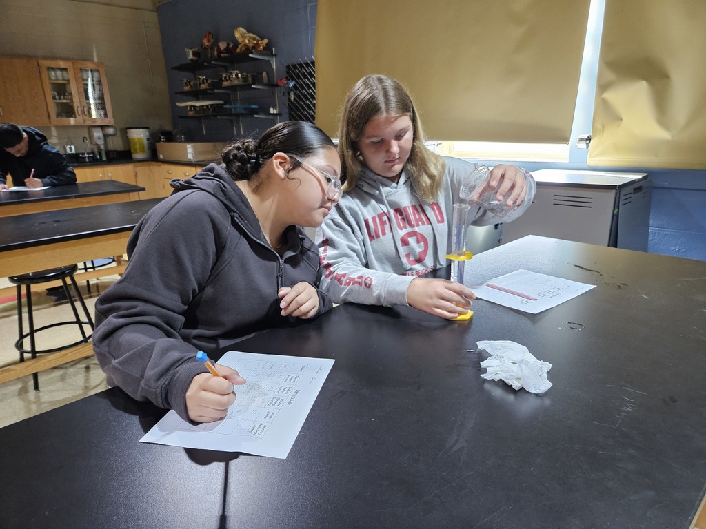 A student pours water into a graduated cylinder to measure while her lab partner observes.