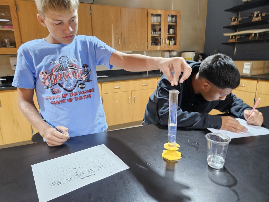 One student drops something into a graduated cylinder with water while his lab partner takes notes.