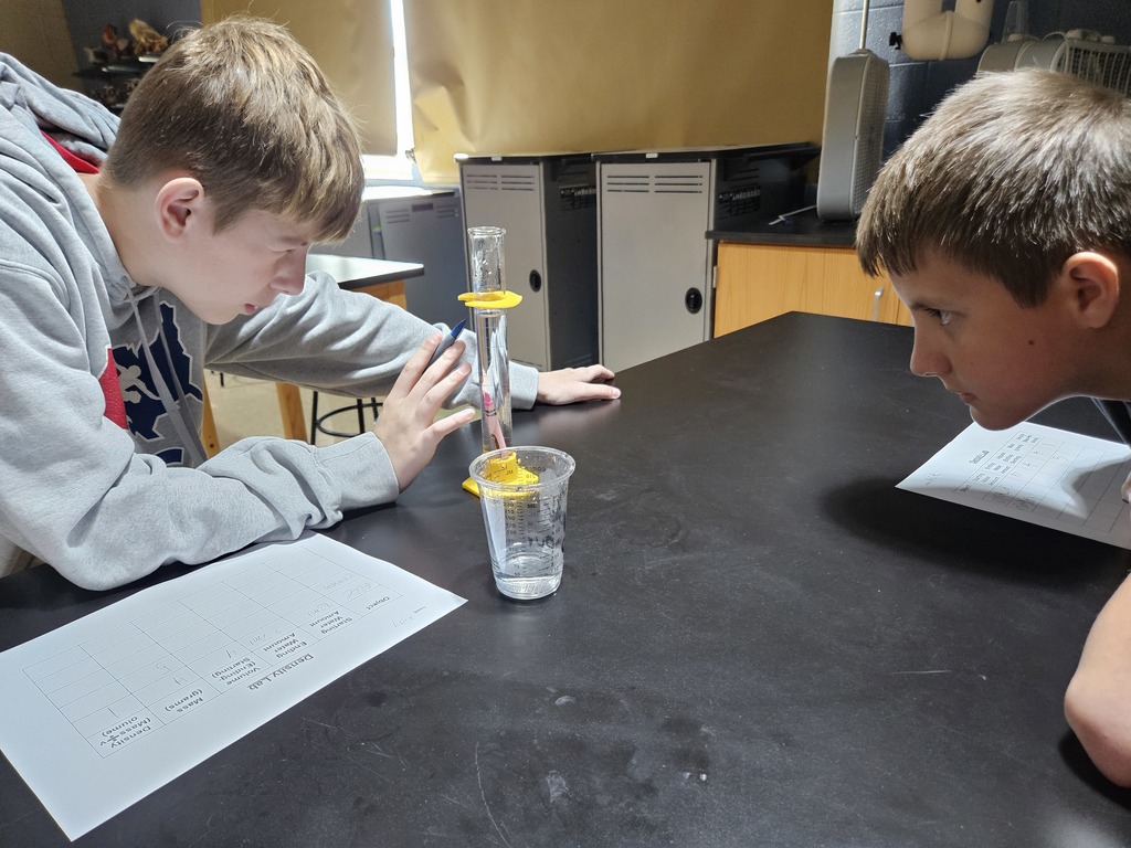 Students lean to observe water displacement of a crayon in a graduated cylinder to measure volume.