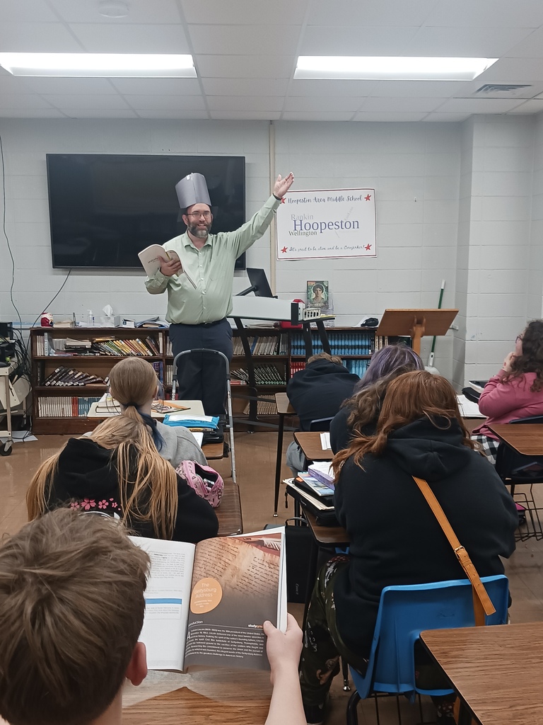 Teacher standing on step stool with construction paper top hat, pretending to be Abraham Lincoln delivering the Gettysburg Address.